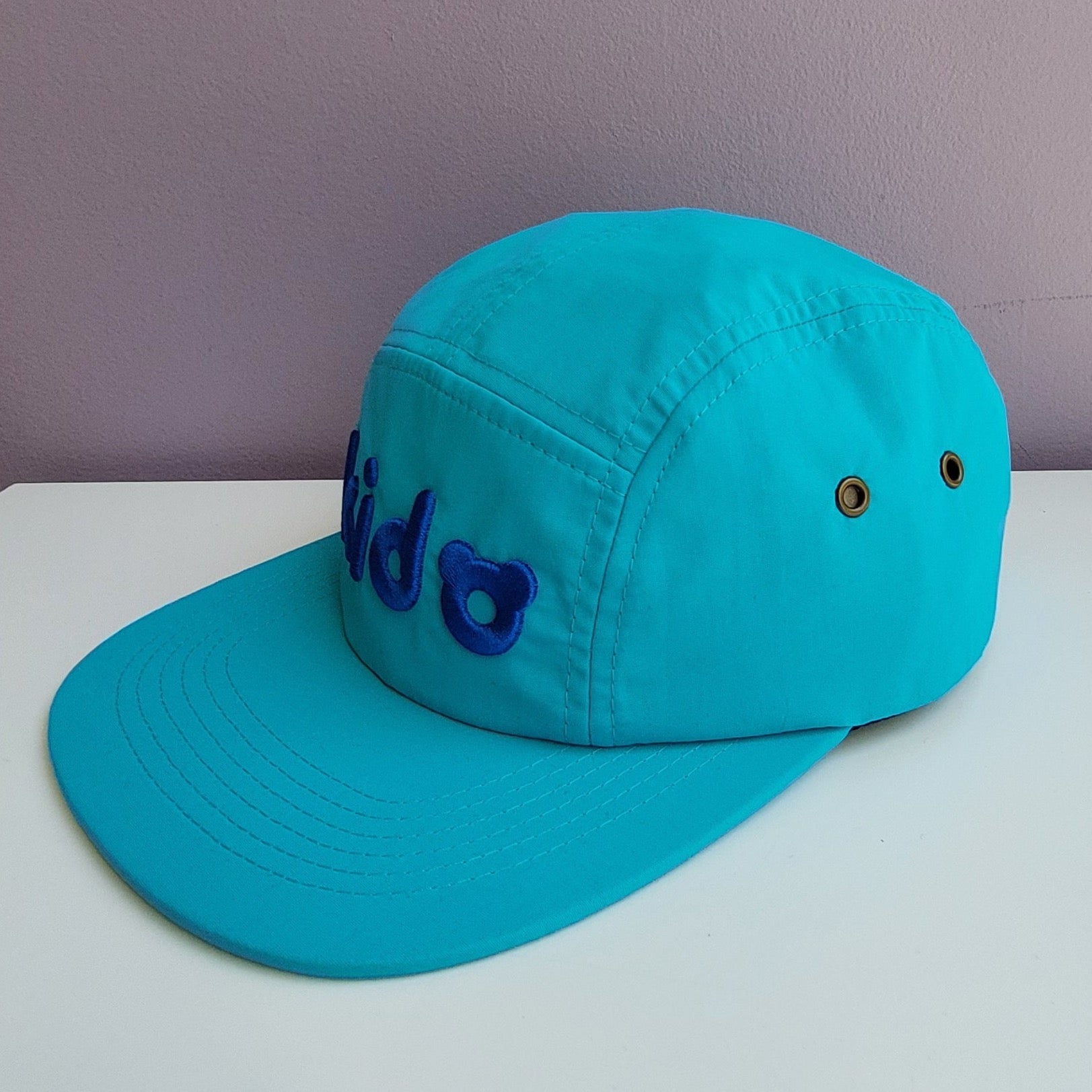 A bright blue 5 panel cap turned at a 45 degree angle sits on a white surface with a light purple background. The Kido logo is embroidered across the front in darker blue thread.