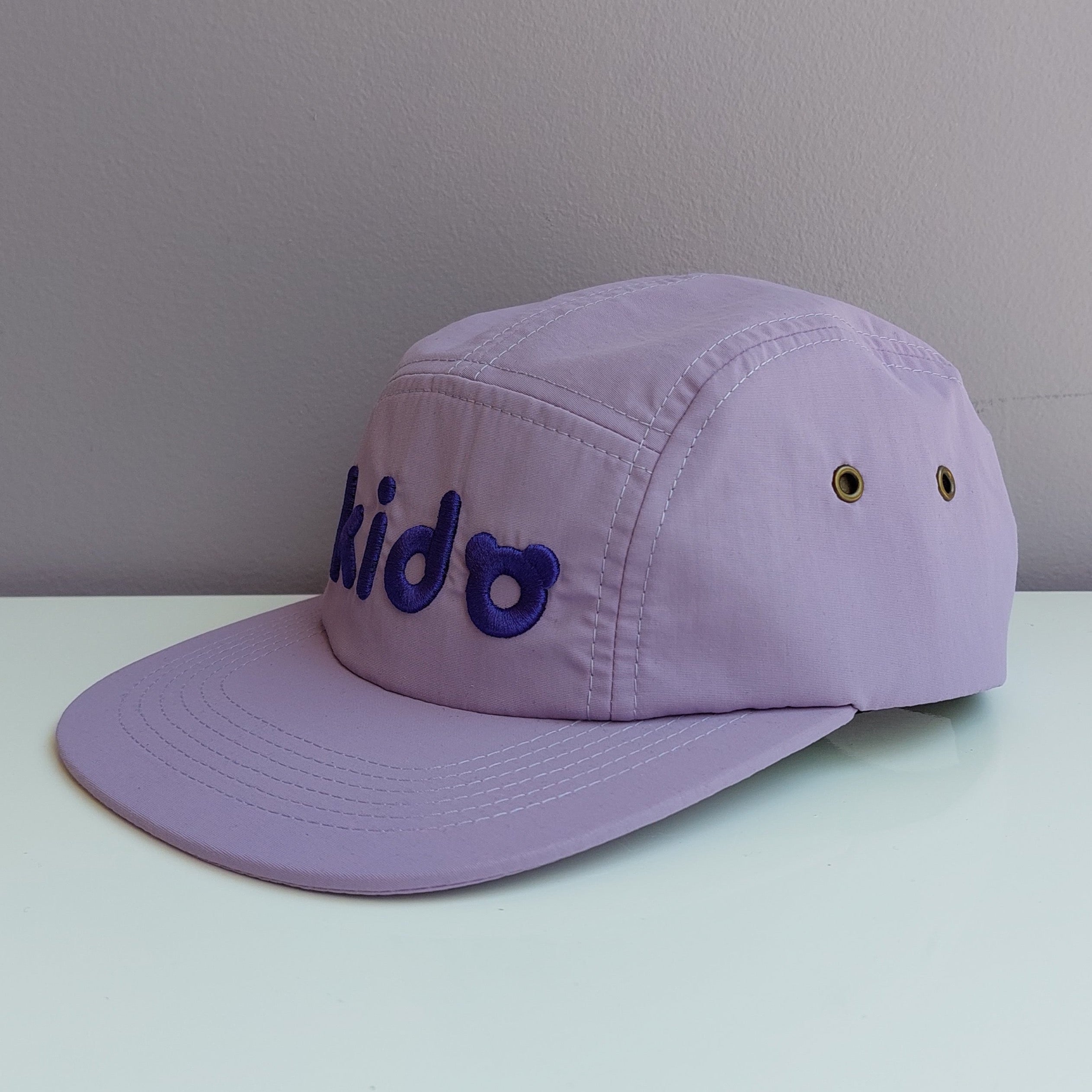 A light purple 5 panel cap turned at a 45 degree angle sits on a white surface with a light purple background. The Kido logo is embroidered across the front in darker purple thread.