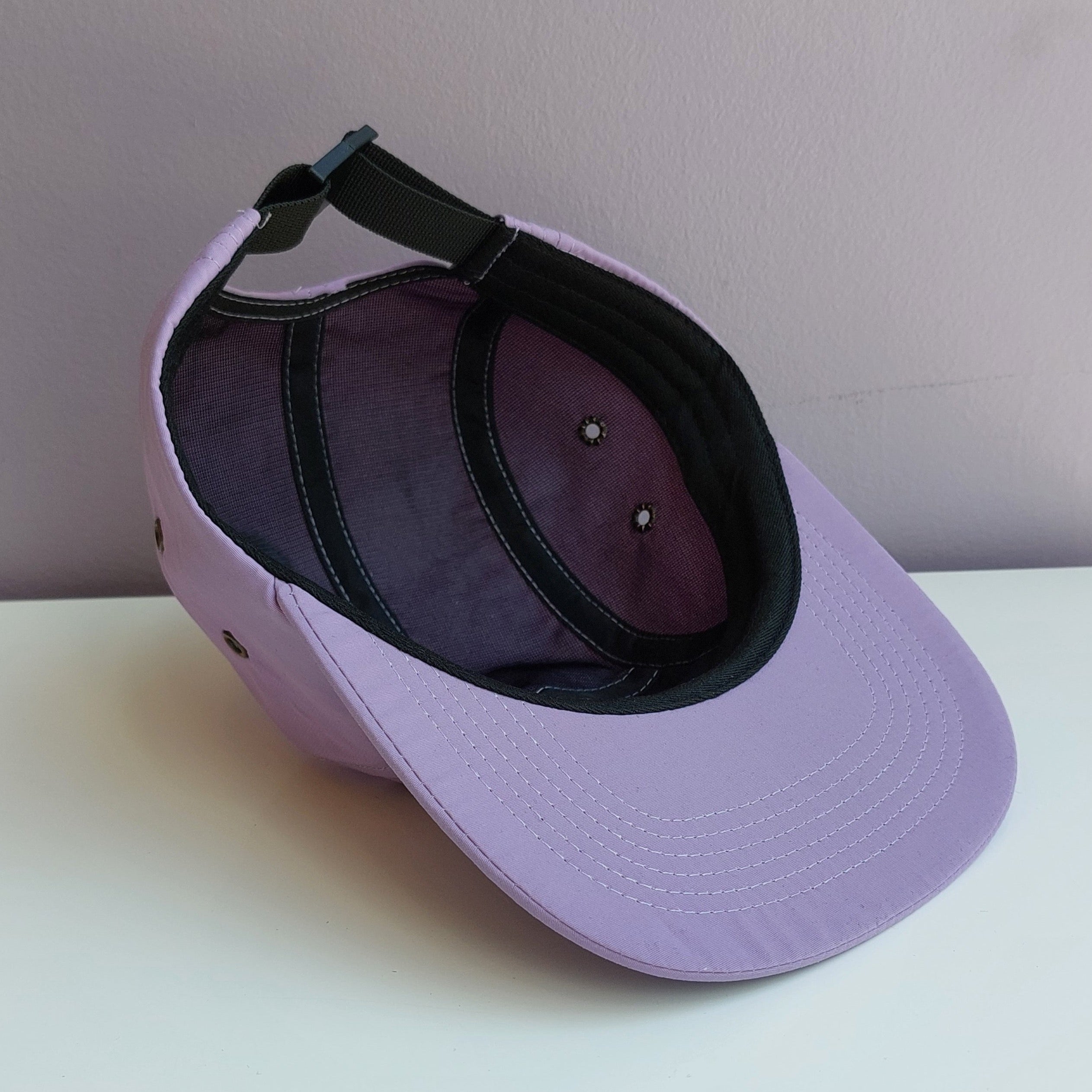 A light purple 5 panel cap with black mesh interior is upside down & turned at a 45 degree angle sitting on a white surface with a light purple background.