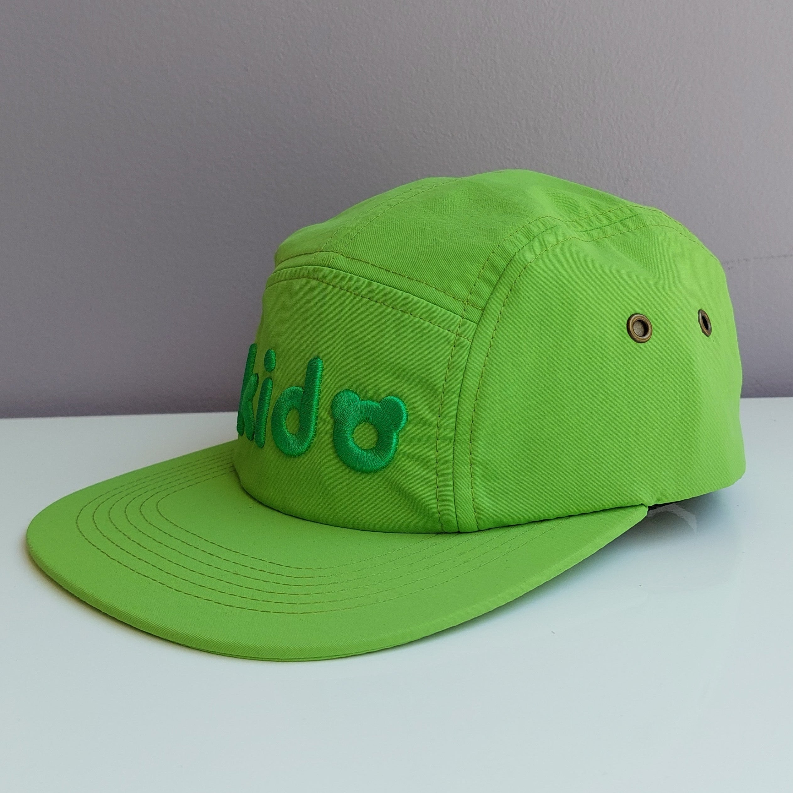 A bright green 5 panel cap turned at a 45 degree angle sits on a white surface with a light purple background. The Kido logo is visible across the front in darker green embroidery.