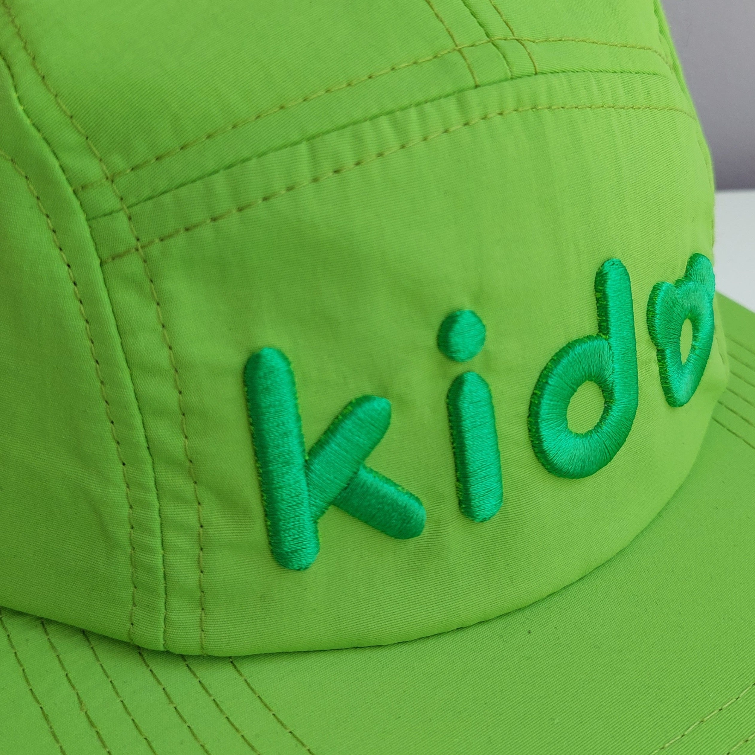 A close up of a bright green 5 panel cap turned at a 45 degree angle sits on a white surface with a light purple background. The Kido logo is visible across the front in darker green embroidery.