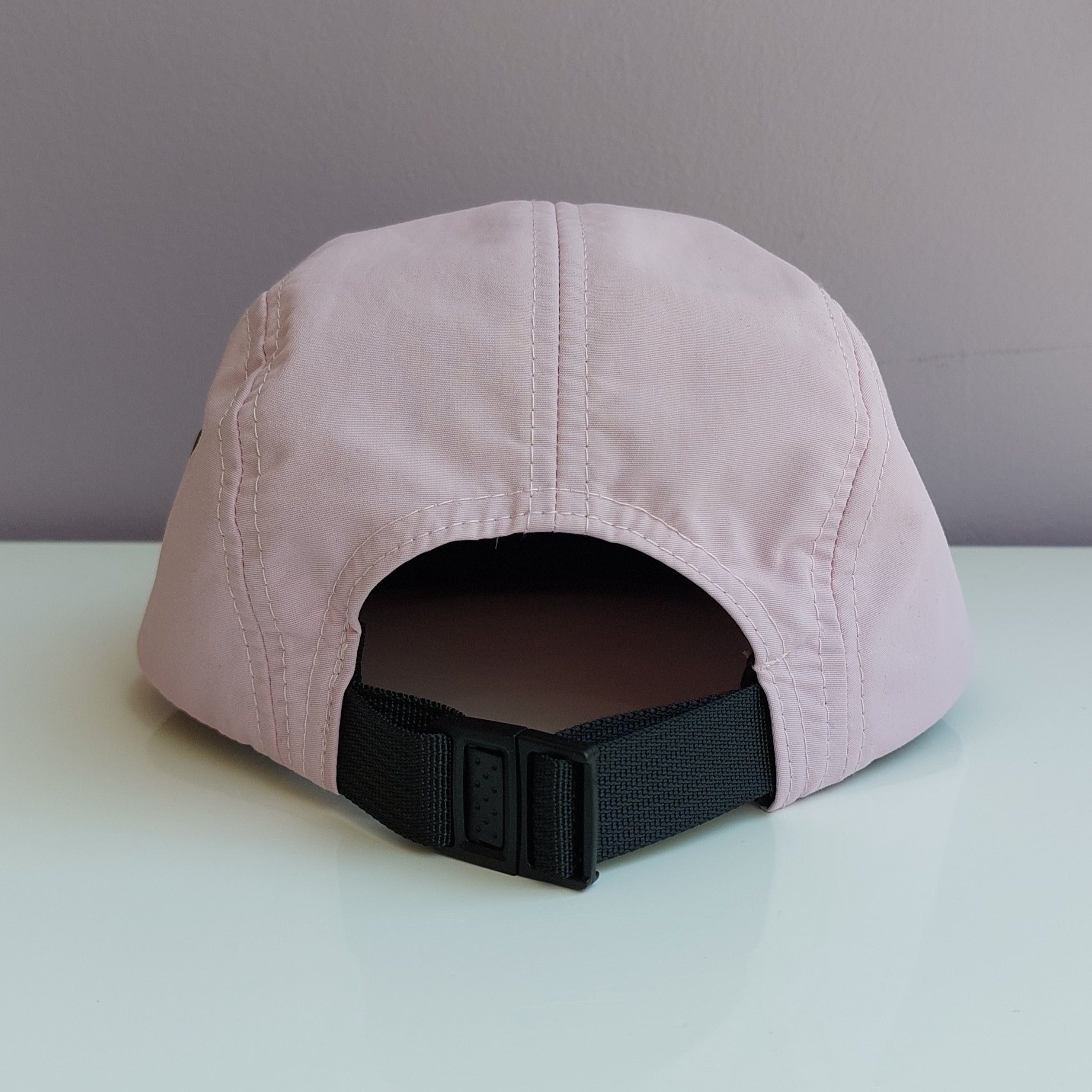 A back view of a pale pink 5 panel cap with a black adjustable strap sitting on a white surface with a light purple background.