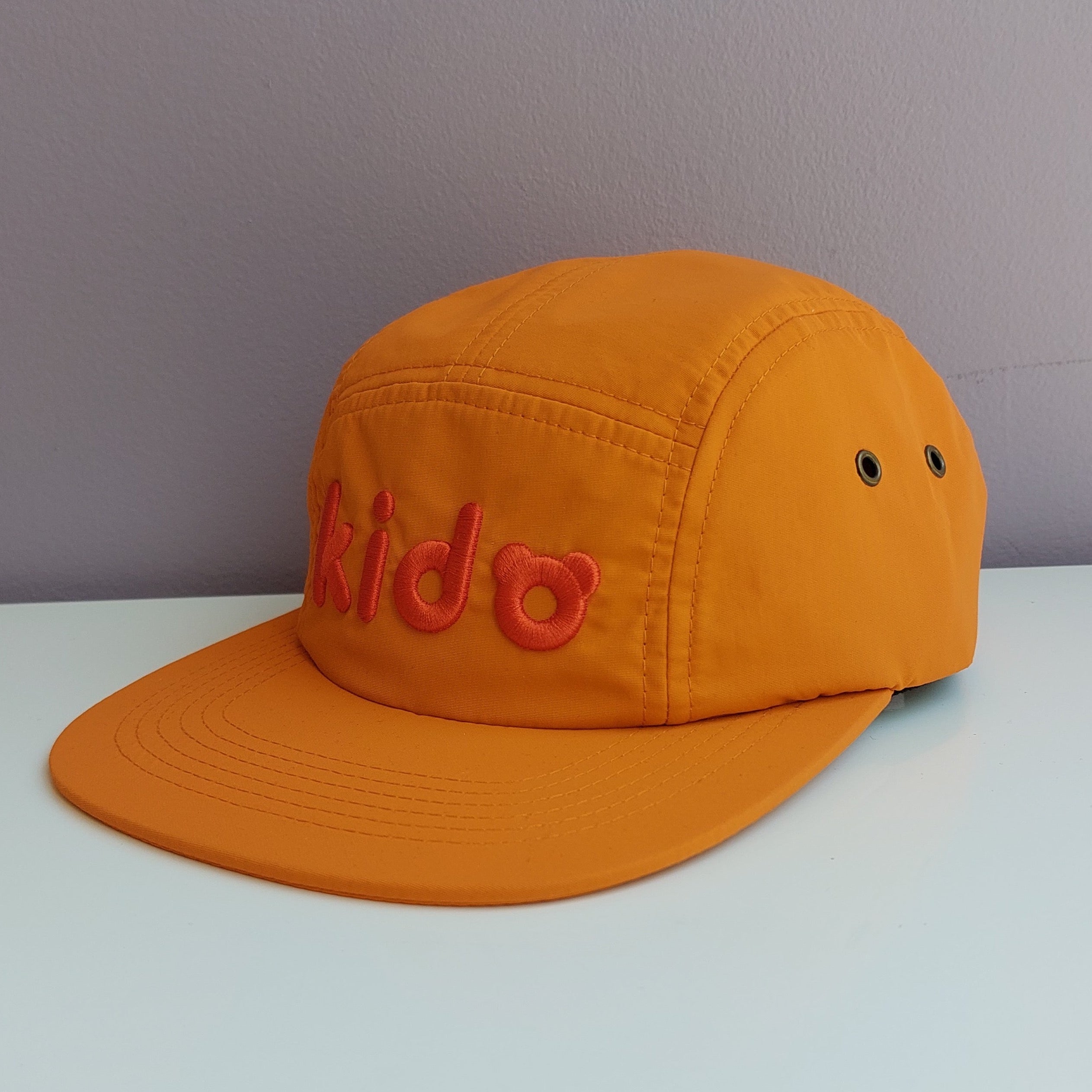 An orange 5 panel cap turned at a 45 degree angle sits on a white surface with a light purple background. The Kido logo is embroidered across the front in slightly darker orange thread.
