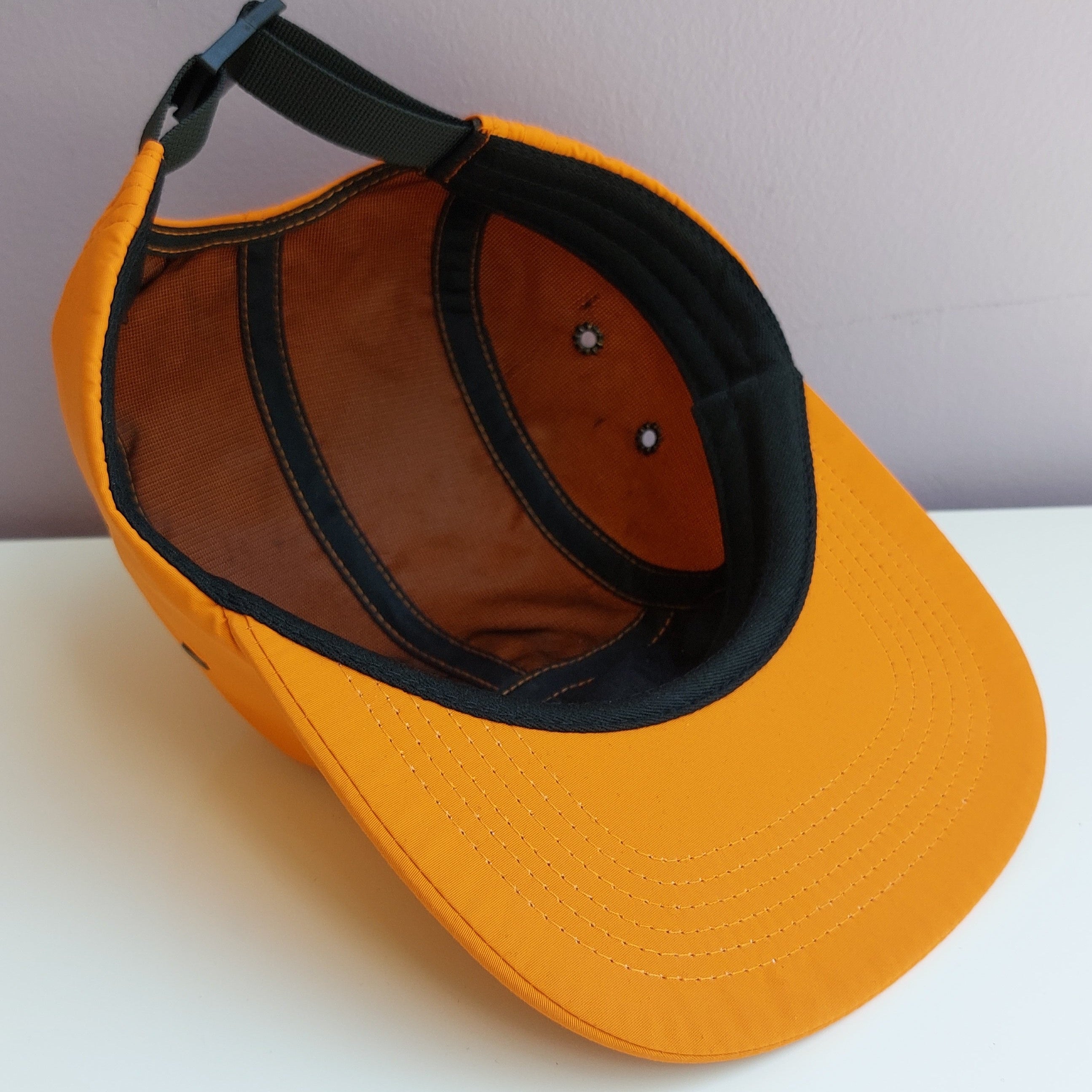 An orange 5 panel cap with black mesh interior is upside down & turned at a 45 degree angle sitting on a white surface with a light purple background.