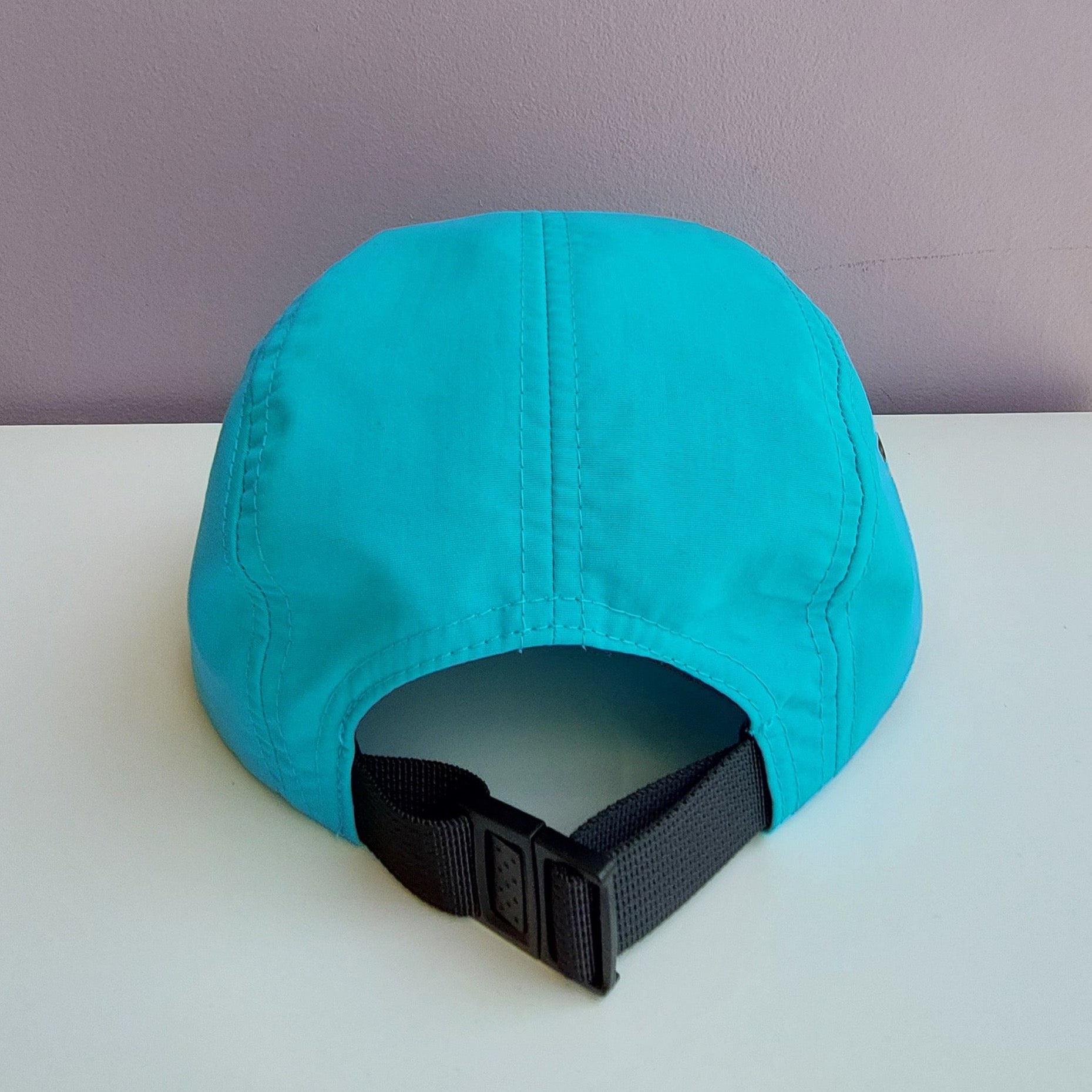 A back view of a bright blue 5 panel cap with a black adjustable strap sitting on a white surface with a light purple background.