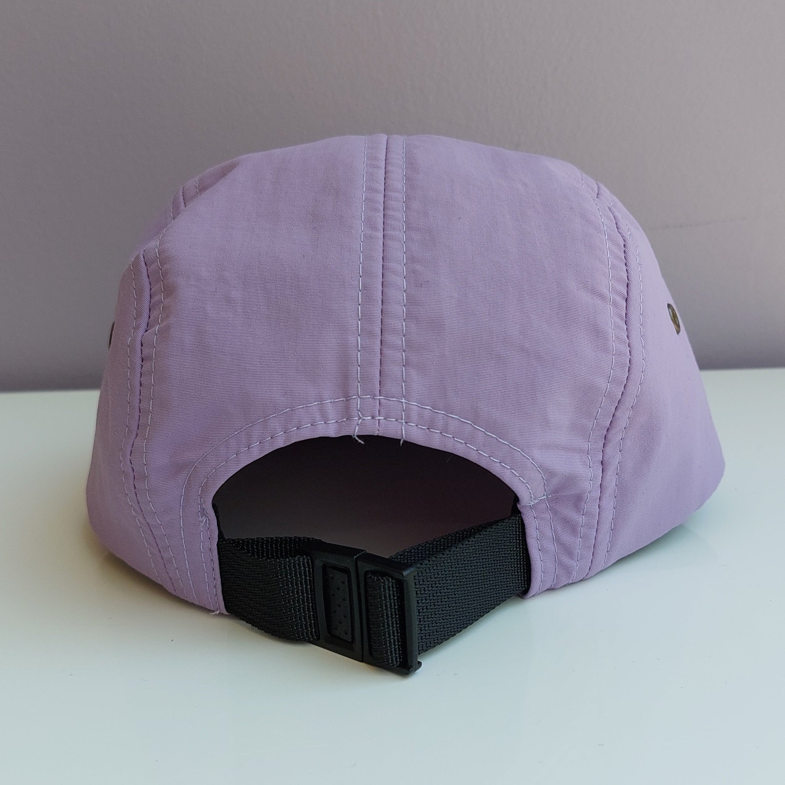 A back view of a light purple 5 panel cap with a black adjustable strap sitting on a white surface with a light purple background.