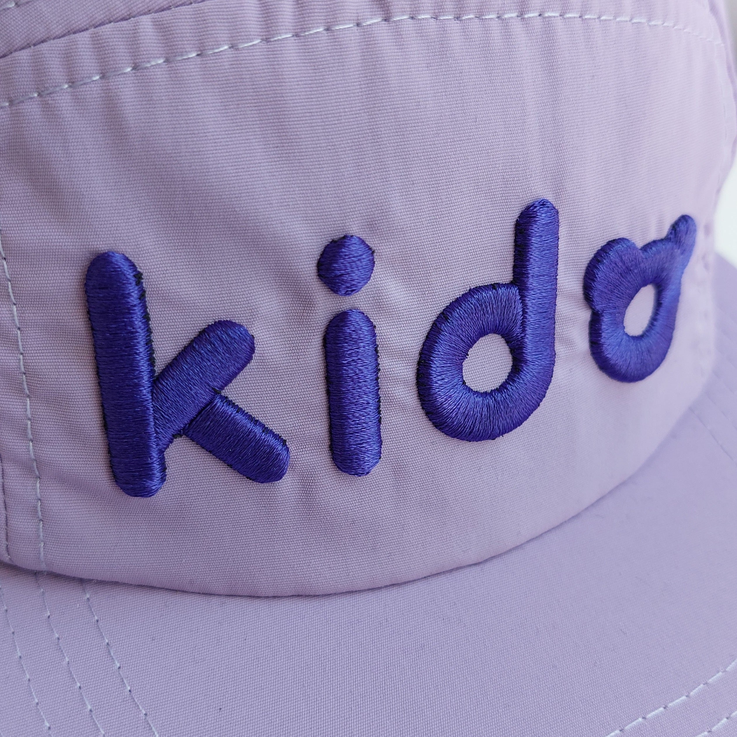 A close up of a light purple 5 panel cap turned at a 45 degree angle sits on a white surface with a light purple background. The Kido logo is embroidered across the front in darker purple thread.