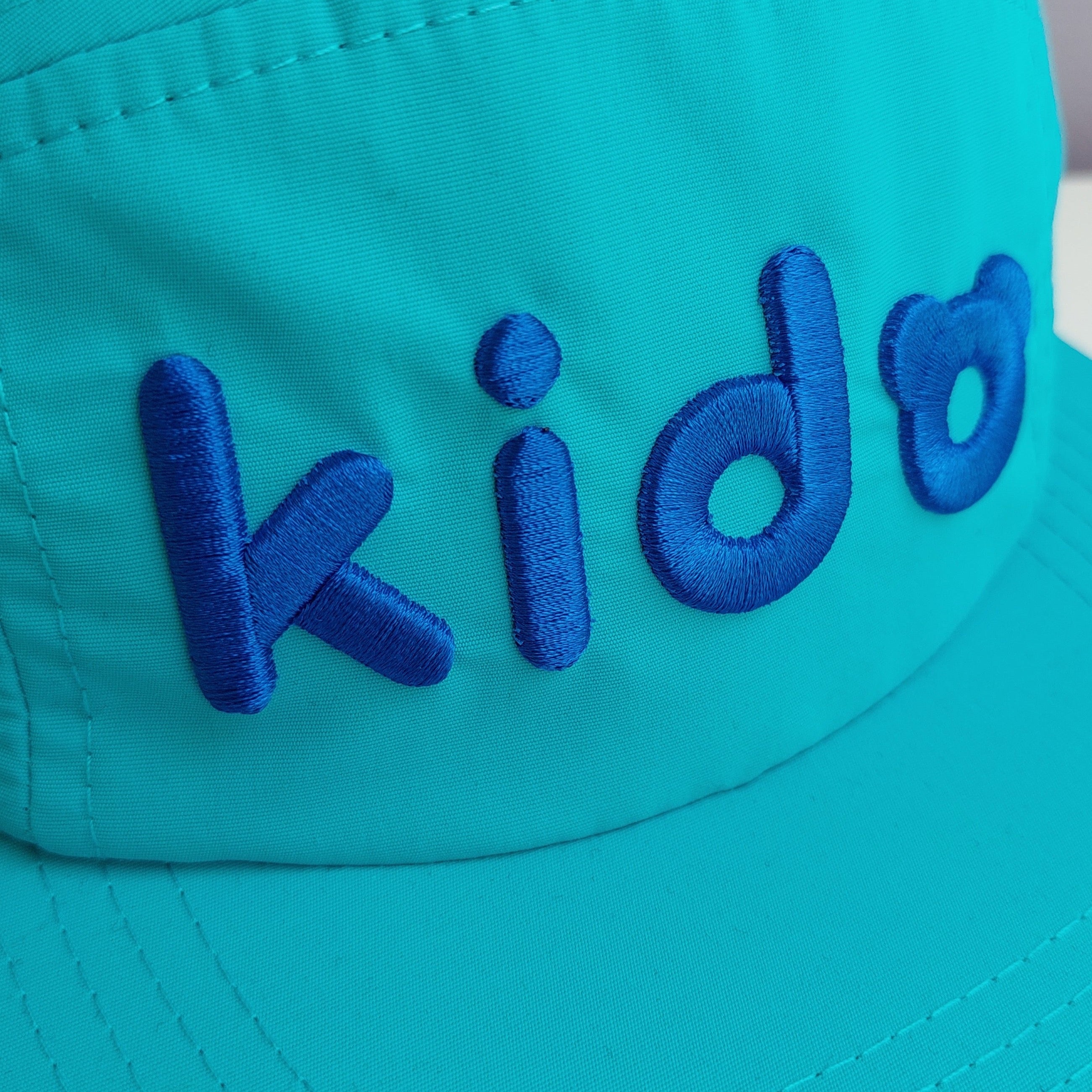 A close up of A bright blue 5 panel cap turned at a 45 degree angle sits on a white surface with a light purple background. The Kido logo is embroidered across the front in darker blue thread.