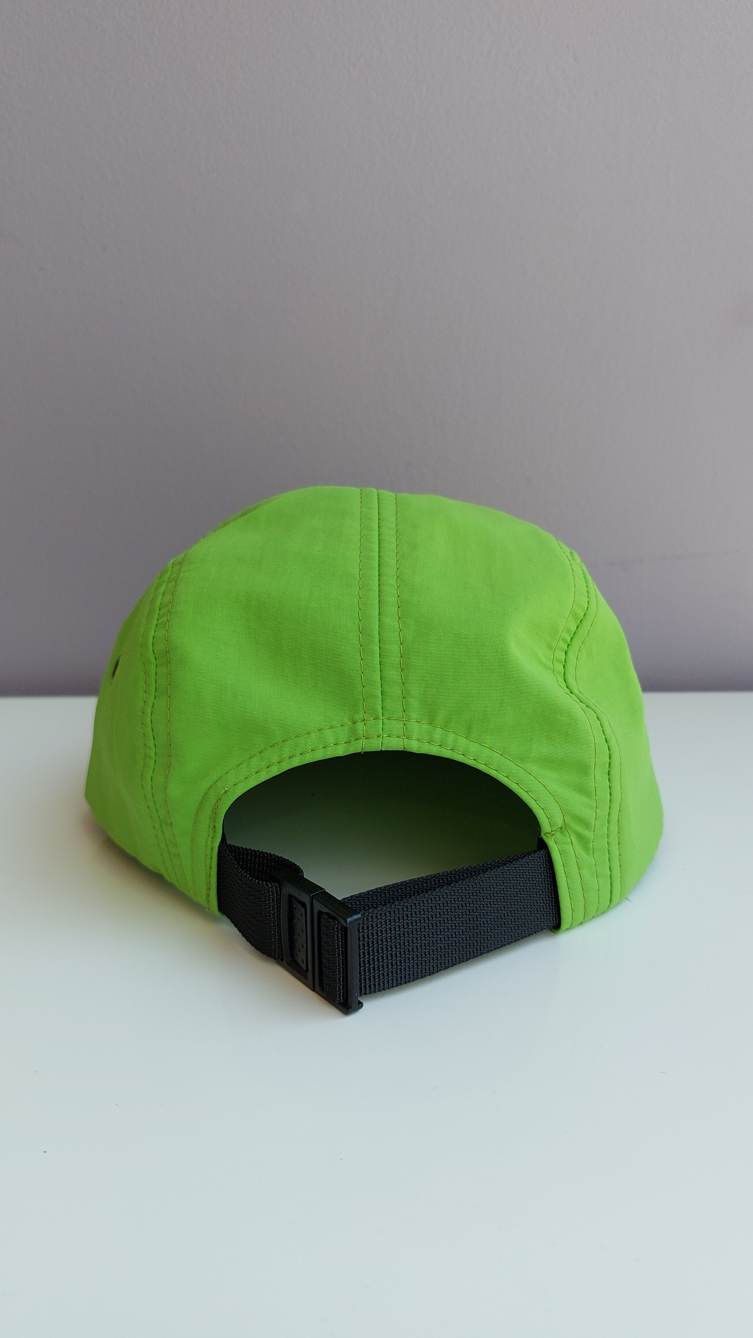 A back view of a bright green 5 panel cap with a black adjustable strap sitting on a white surface with a light purple background.