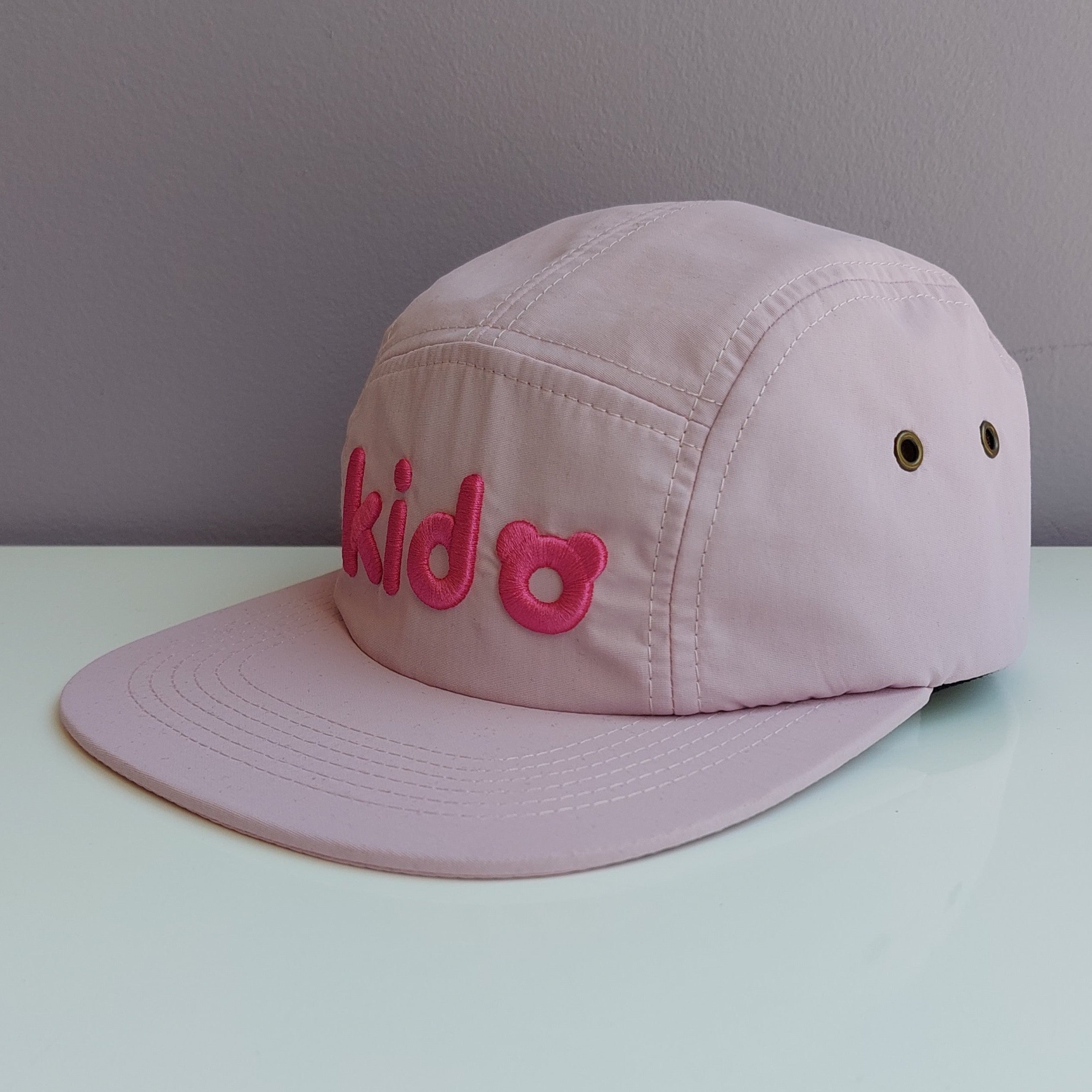 A pale pink 5 panel cap turned at a 45 degree angle sits on a white surface with a light purple background. The Kido logo is embroidered across the front in darker pink thread.