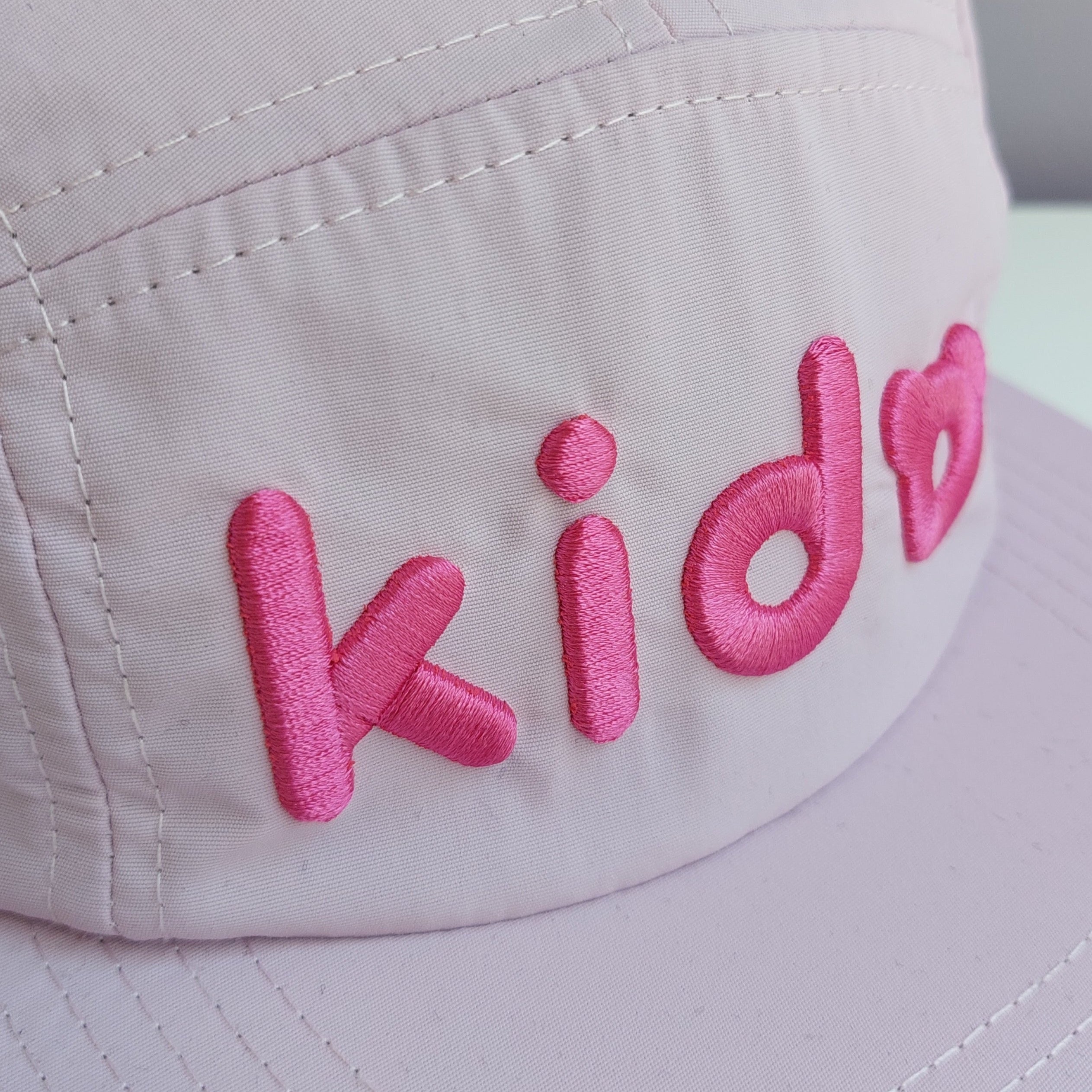 A close up of a pale pink 5 panel cap turned at a 45 degree angle sits on a white surface with a light purple background. The Kido logo is embroidered across the front in darker pink thread.