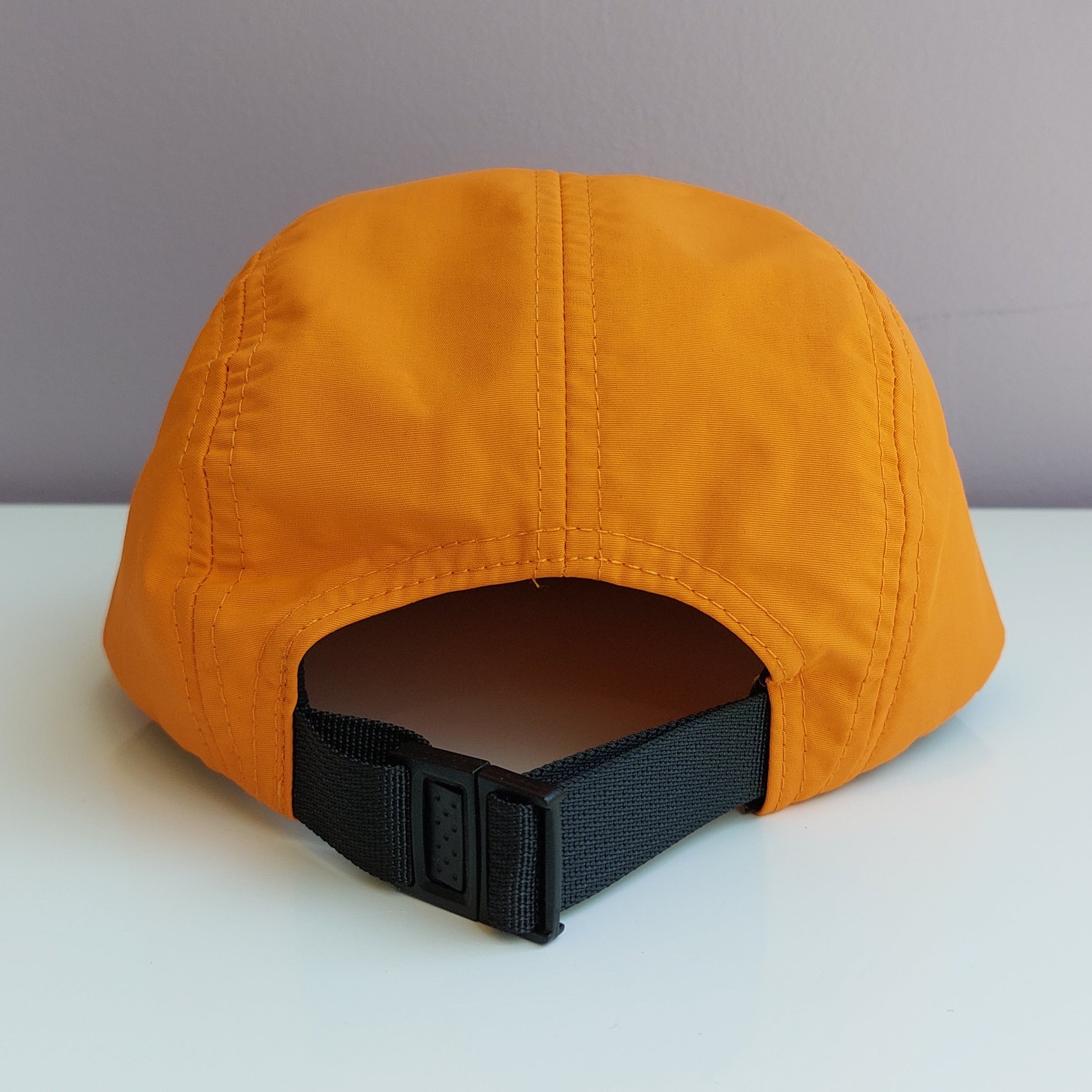 A back view of an orange 5 panel cap with a black adjustable strap sitting on a white surface with a light purple background.