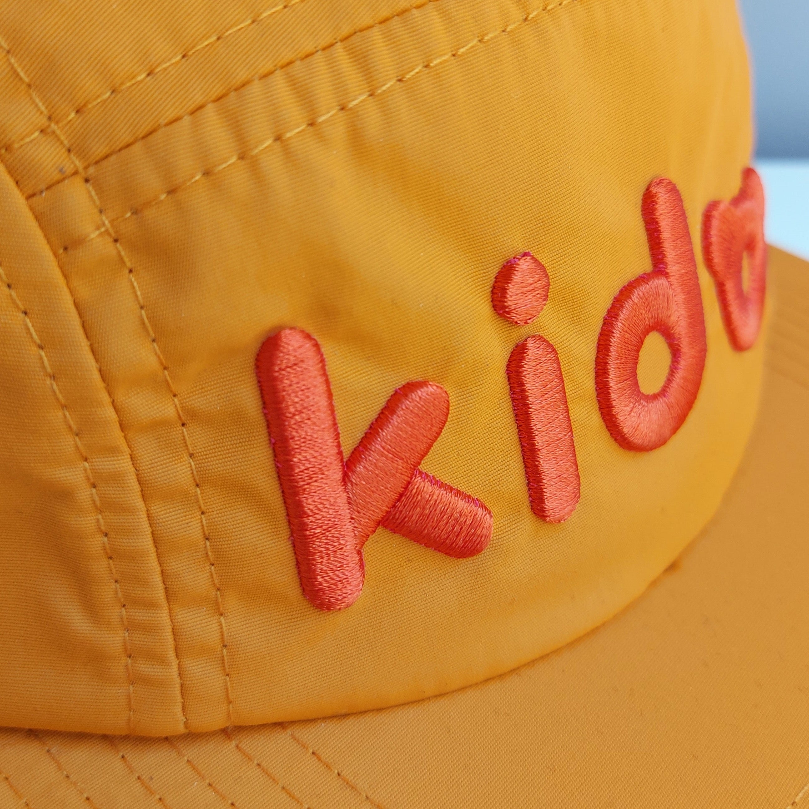 A close up of an orange 5 panel cap turned at a 45 degree angle sits on a white surface with a light purple background. The Kido logo is embroidered across the front in slightly darker orange thread.