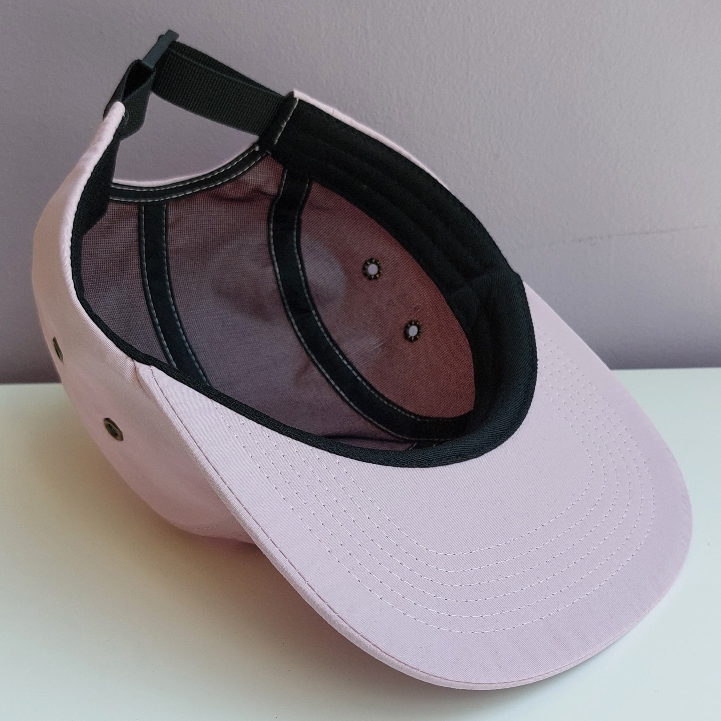 A pale pink 5 panel cap with black mesh interior is upside down & turned at a 45 degree angle sitting on a white surface with a light purple background.