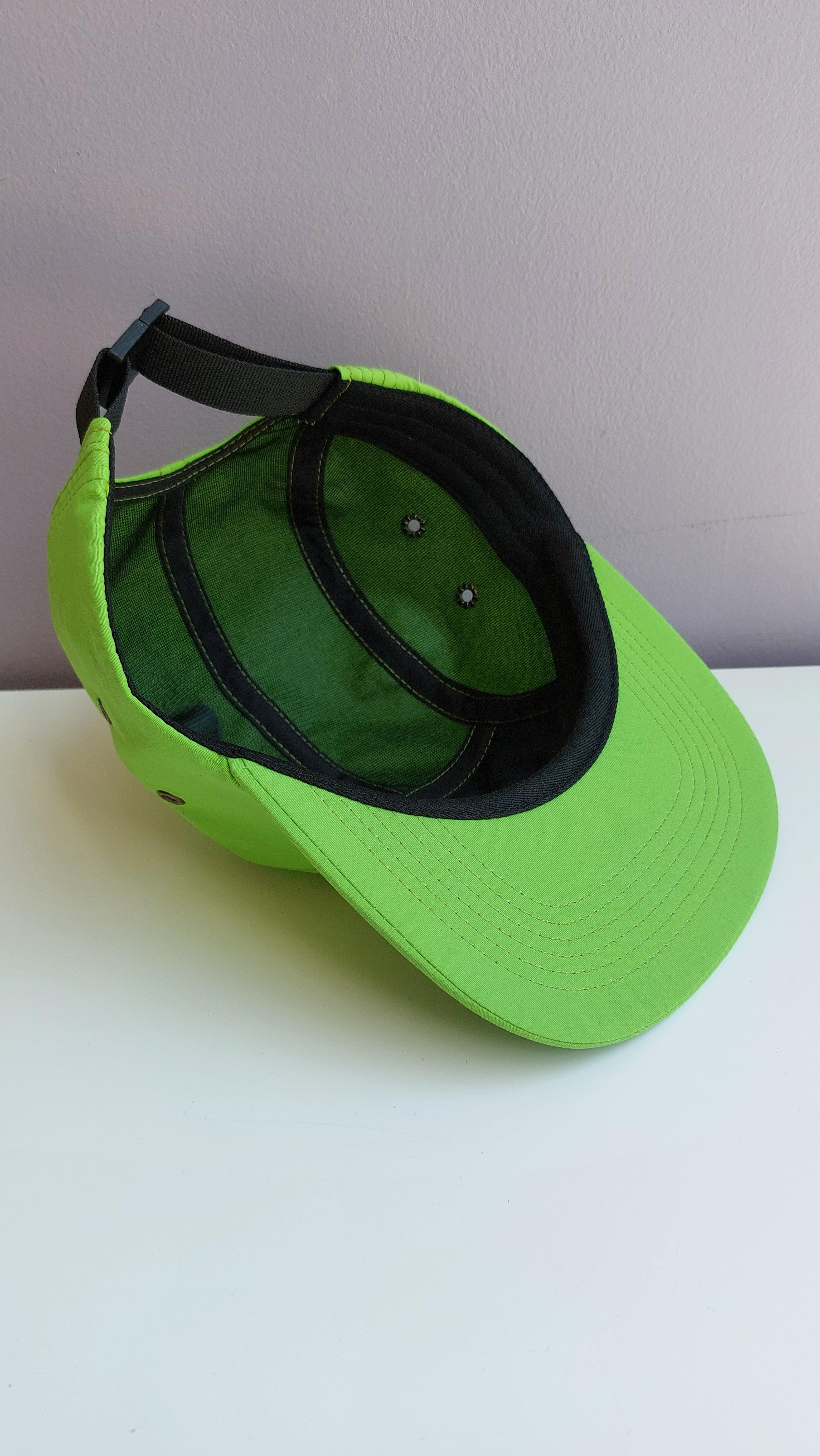 A bright green 5 panel cap with black mesh interior is upside down & turned at a 45 degree angle sitting on a white surface with a light purple background.