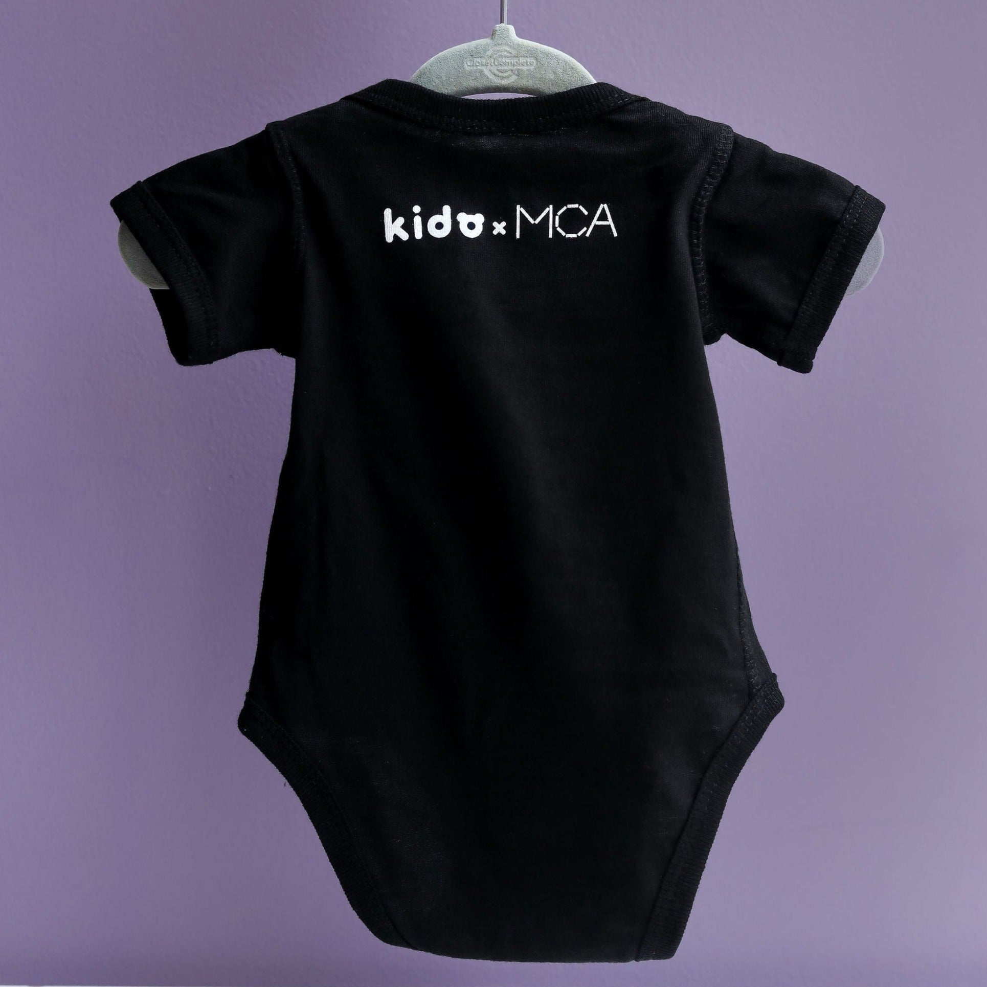 back of a black onesie on a purple background white text, printed at the nape of the neck is kido x MCA